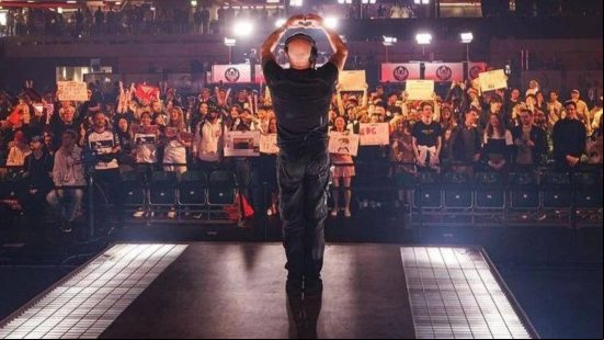 A bald man in front of a crowd wearing headphones forms a heart with his hands as they cheer and hold up signs